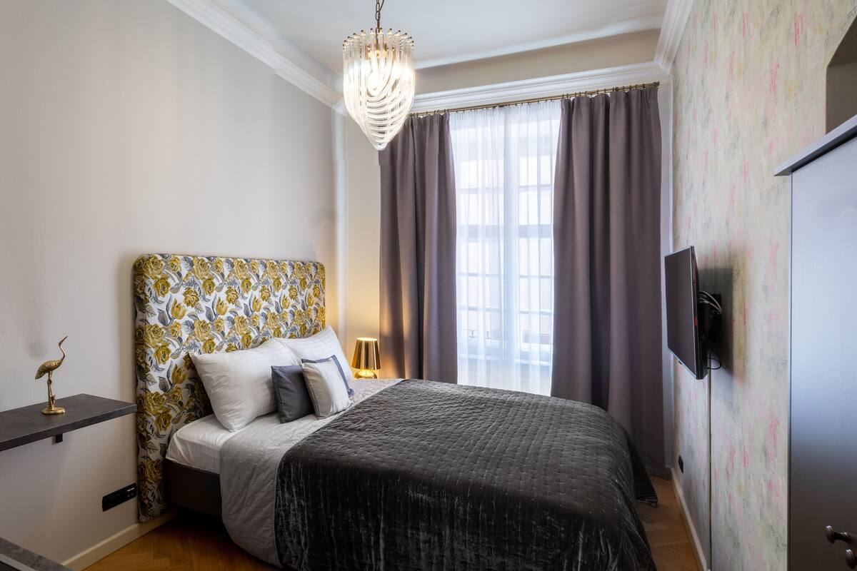 Un dormitorio con cabecera estampada, cama con colcha oscura, lámpara de noche, televisor montado en la pared y ventana con cortinas.