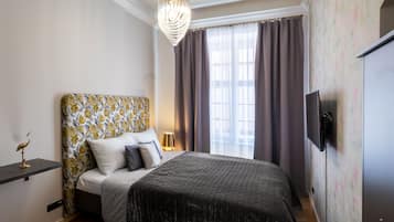 Een slaapkamer met een bekleed hoofdeinde, een bed met een donge deken, een bedlampje, een aan de muur bevestigde televisie, en een raam met gordijnen.