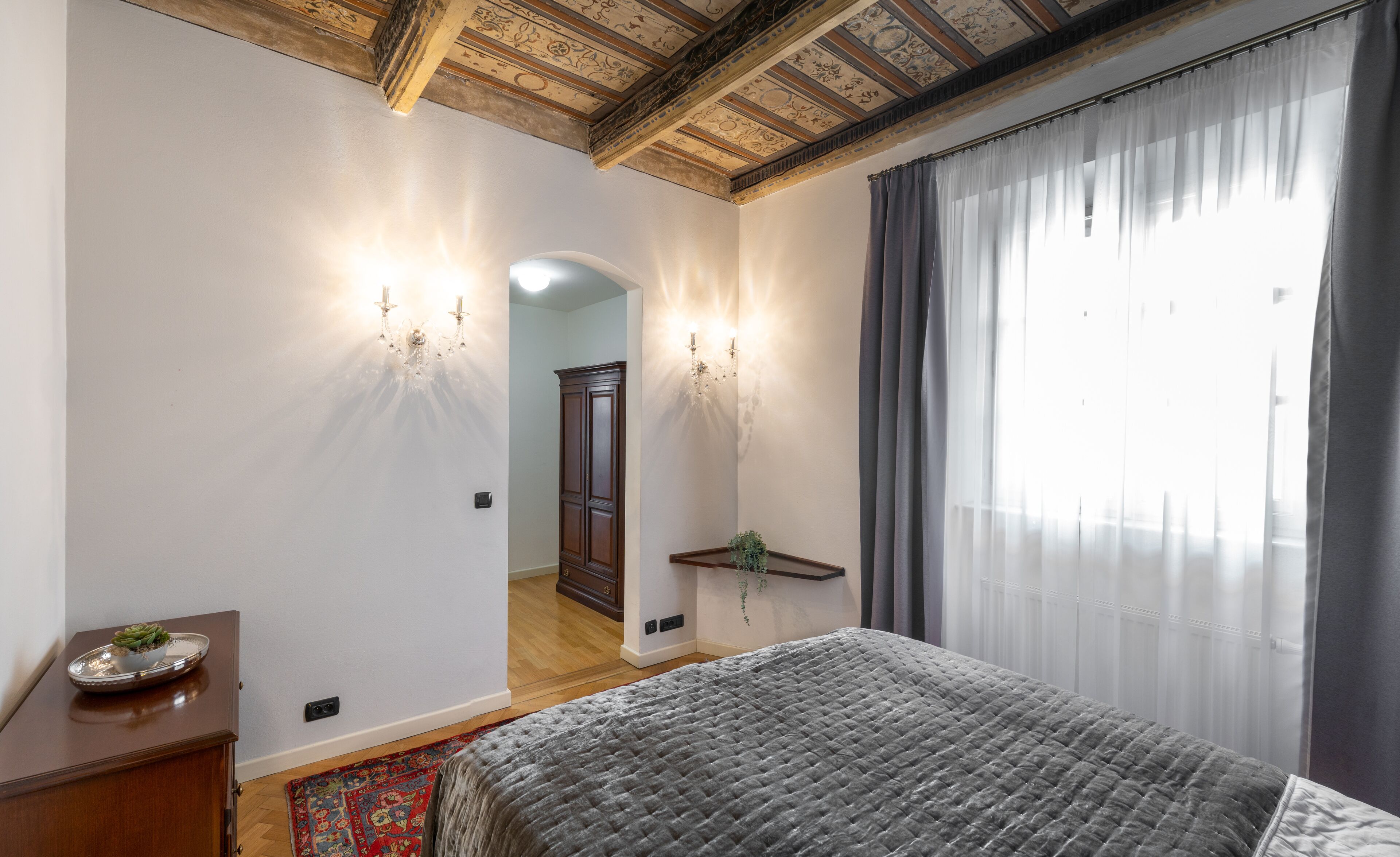 Een slaapkamer met een bed, nachtkastje, dressoir, spiegel en een kroonluchter.