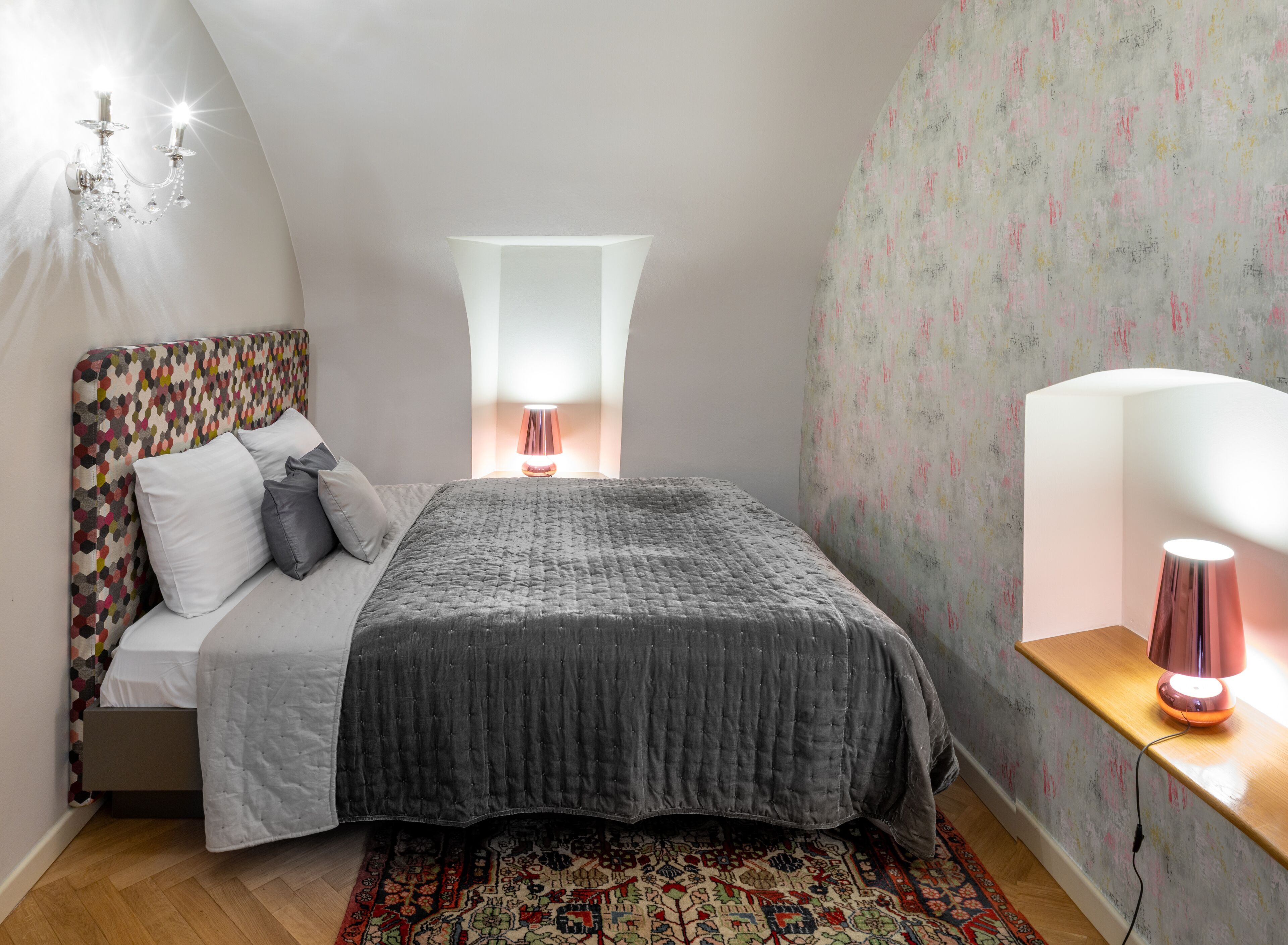 Ein Schlafzimmer mit Bett, Nachttischlampe, Wandleuchte, einem gemusterten Kopfteil und einem Teppich.