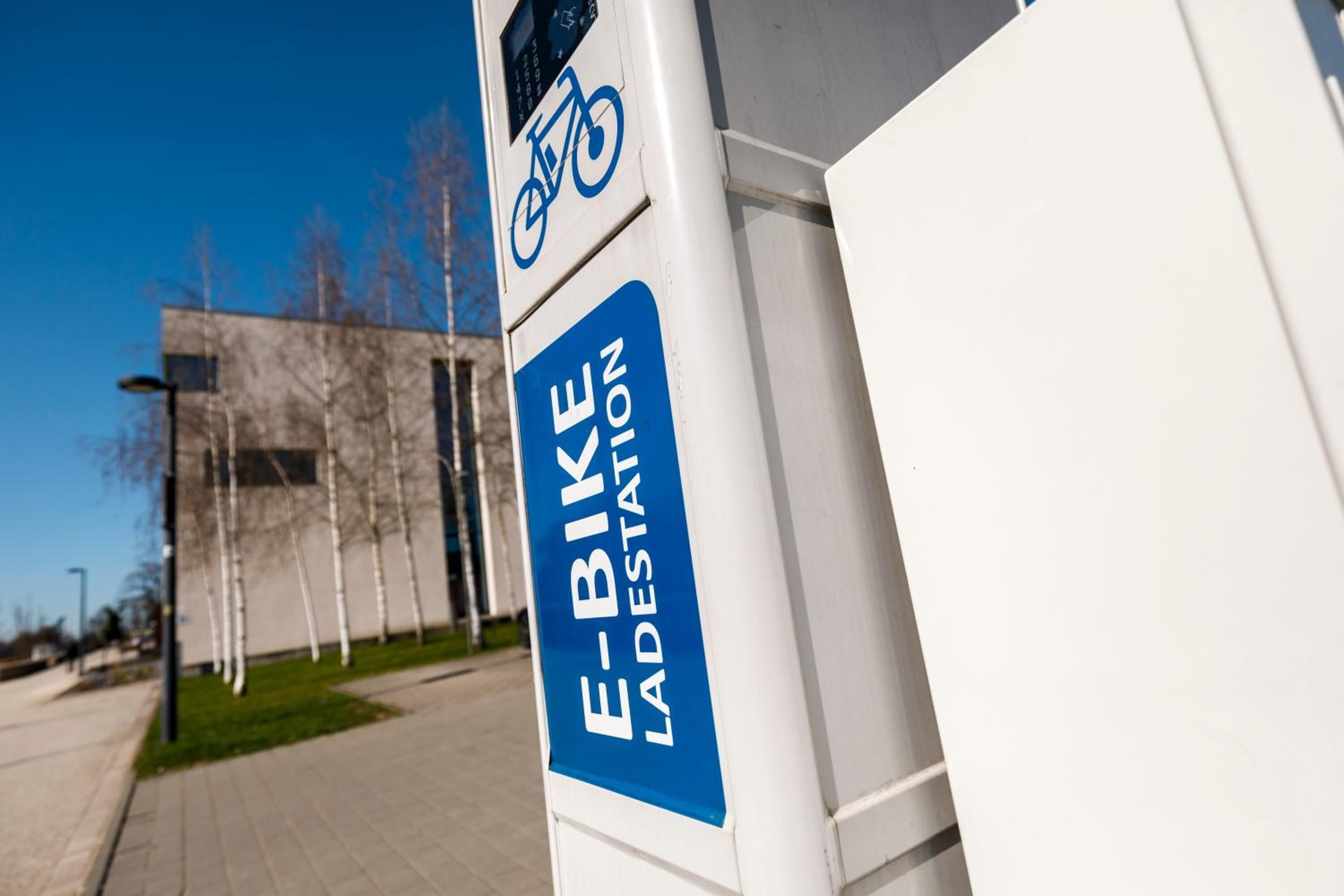 An e-bike station with a clear blue sign, a modern building in the background, and a paved walkway.