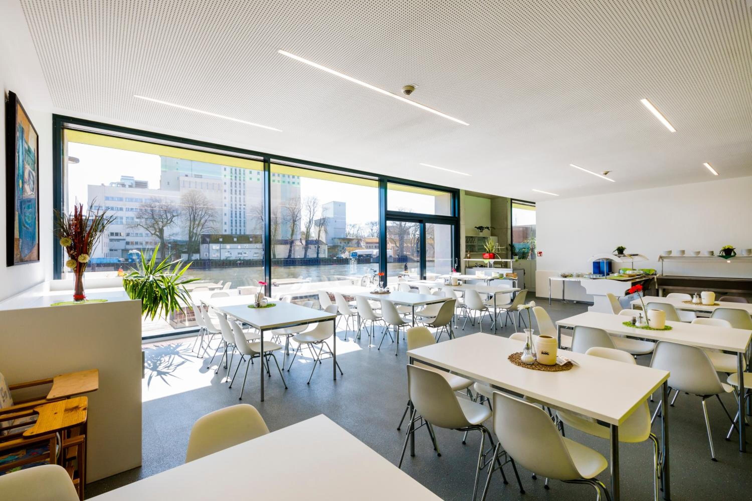A modern dining area with white tables and chairs, large windows offering a city view, and a kitchen area with white cabinets.