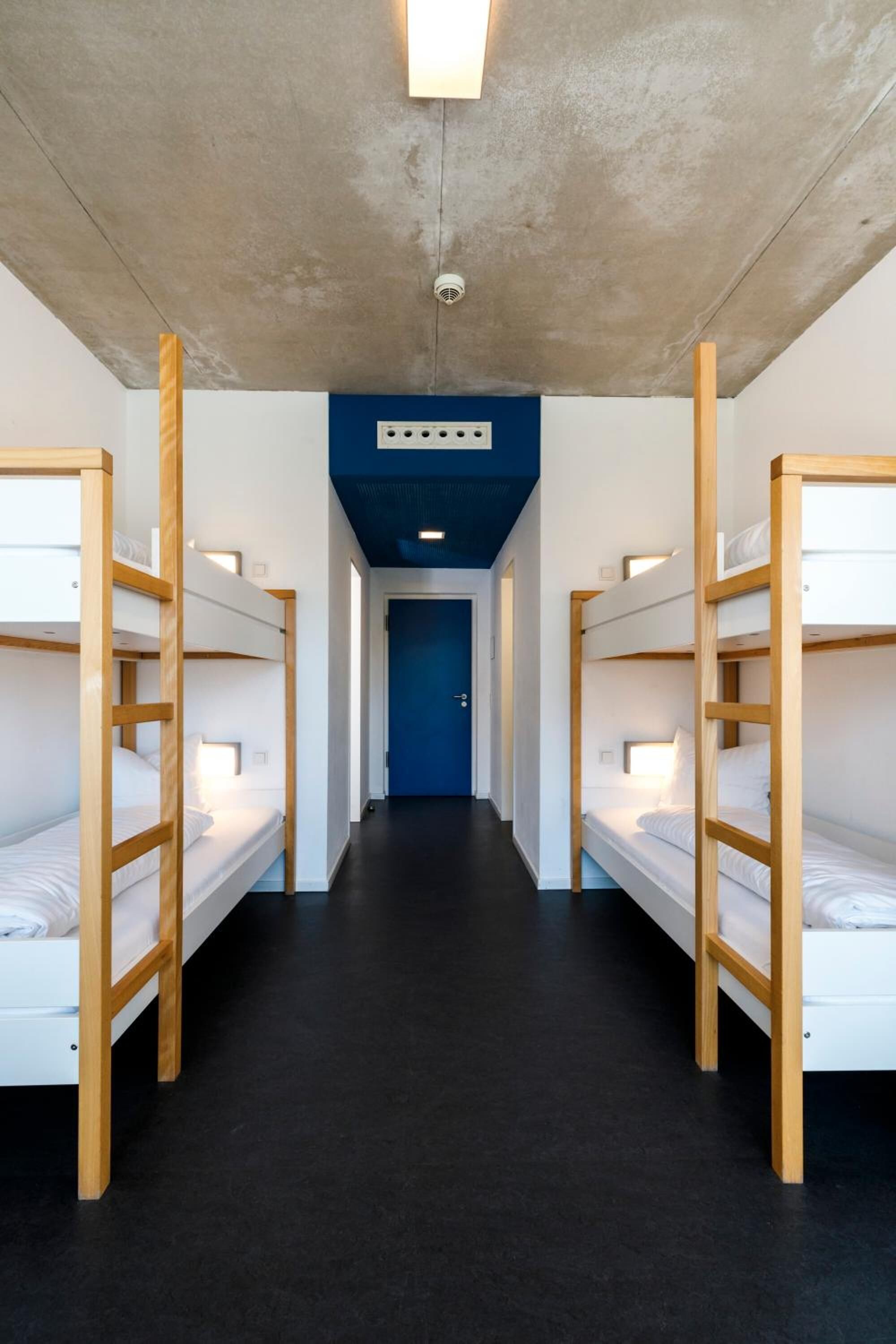 Ein Korridor mit Etagenbetten an beiden Seiten, einer blauen Tür am Ende und einer Deckenleuchte.