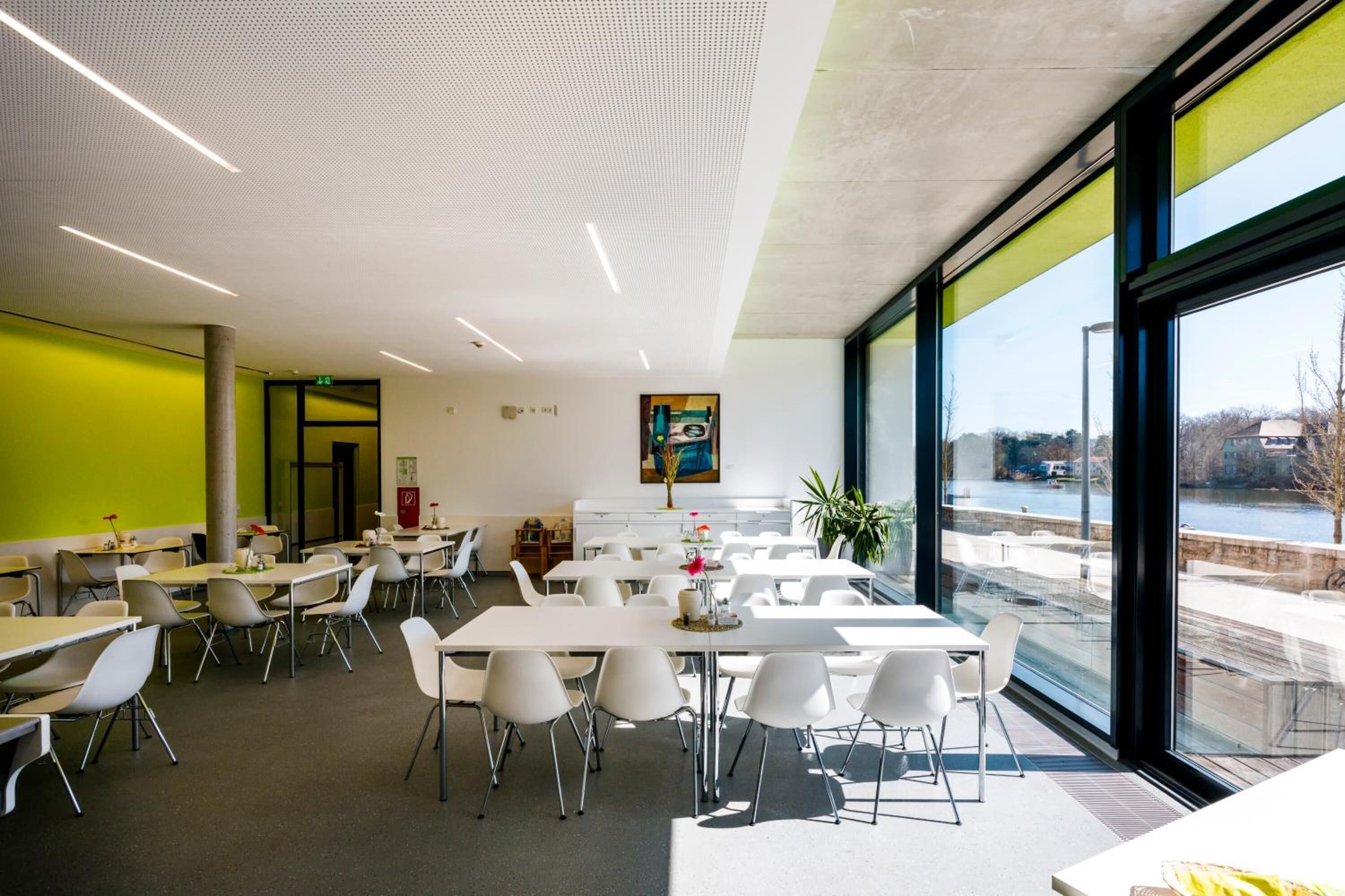 A modern dining area with white tables and chairs, large windows offering a view of the outdoors, and a green accent wall.