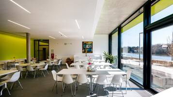 A modern dining area with white tables and chairs, large windows offering a view of the outdoors, and a green accent wall.
