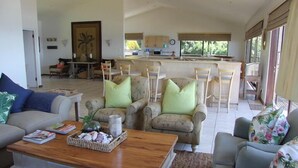 A spacious living room with a sofa, armchairs, and a coffee table. A kitchen area with a dining table and chairs is visible in the background. - Kangela 1 (Ponta d'Ouro)