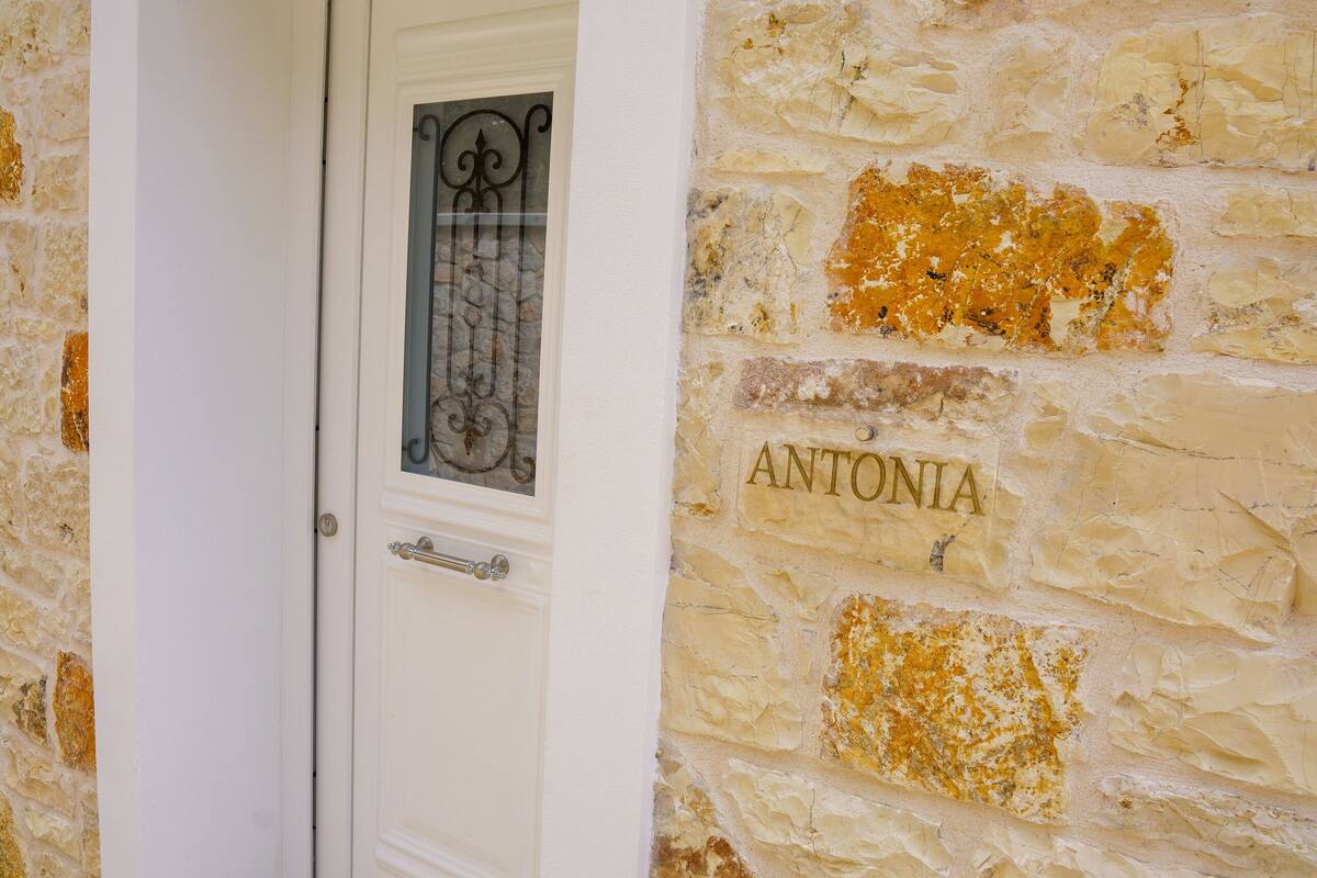 A white door with a glass panel and a metal handle, set in a stone wall with the name "ANTONIA" visible.