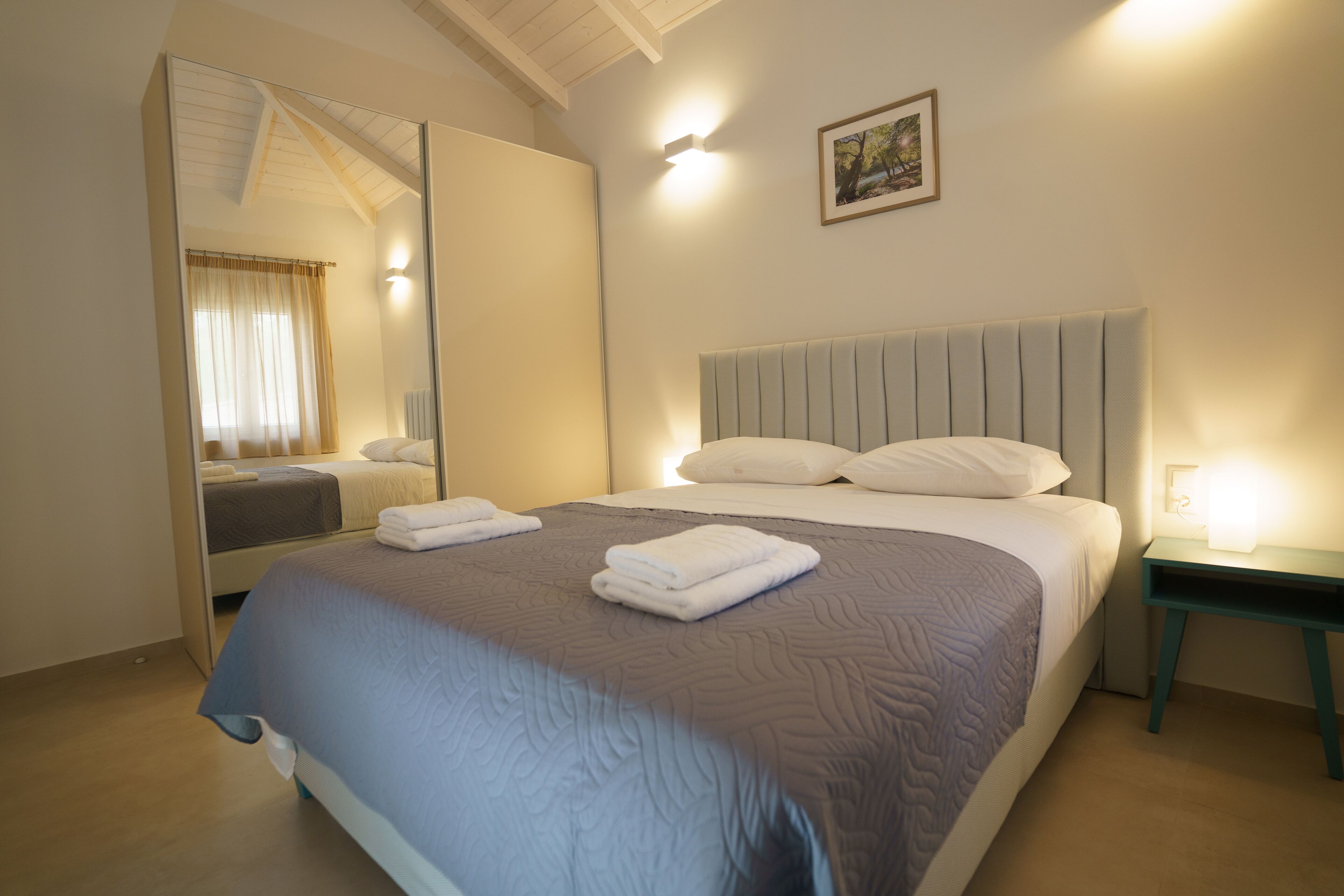 A neatly made bed with white linens and a blue quilt, a bedside table with a lamp, and a large mirror reflecting part of the room.