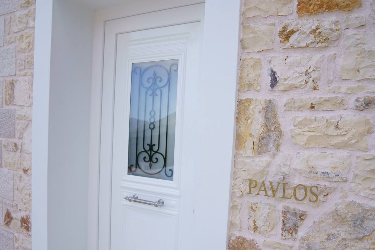 A white door with a glass panel and a metal handle, set in a stone wall with the name "PAVLOS" carved into it.