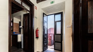 Un pasillo con un extintor fijo en la pared, una puerta que da a un espacio exterior y un suelo de baldosas.