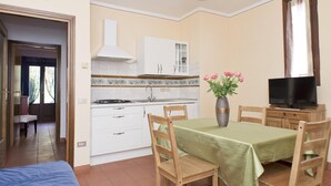 Een compacte keuken met witte kasten, een fornuis en een spoelbak. Er is een eethoek met een tafel en stoelen, en een televisie op een houten dressoir.