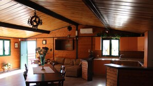 A spacious living area with a wooden ceiling, a sofa, a dining table with chairs, a kitchen area, and a TV mounted on the wall.