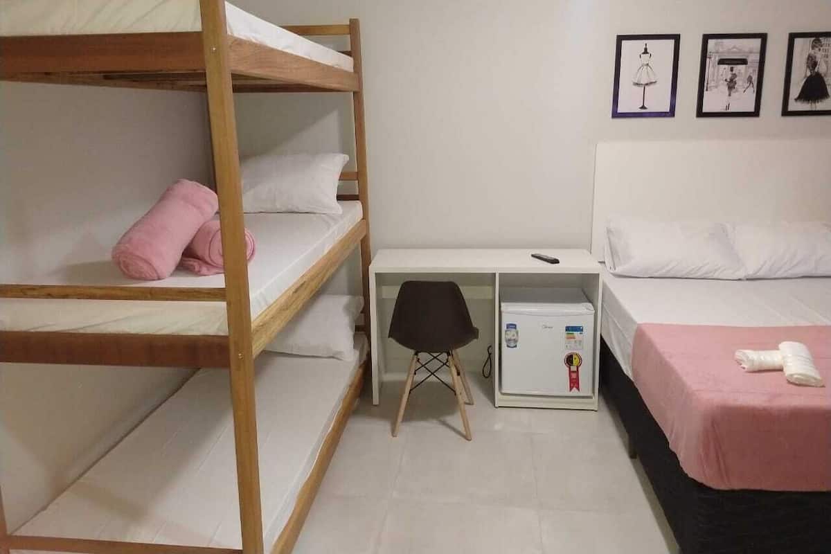 A dormitory room with bunk beds, a desk, a chair, a refrigerator, and a bed with a pink blanket.