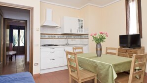 A compact kitchen with white cabinets, a stove, and a sink. There's a dining area with a table and chairs, and a TV on a wooden stand.