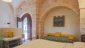 A bedroom with a large bed, a wooden bench, and a small table with folded towels. There is a painting on the wall and an arched stone ceiling.