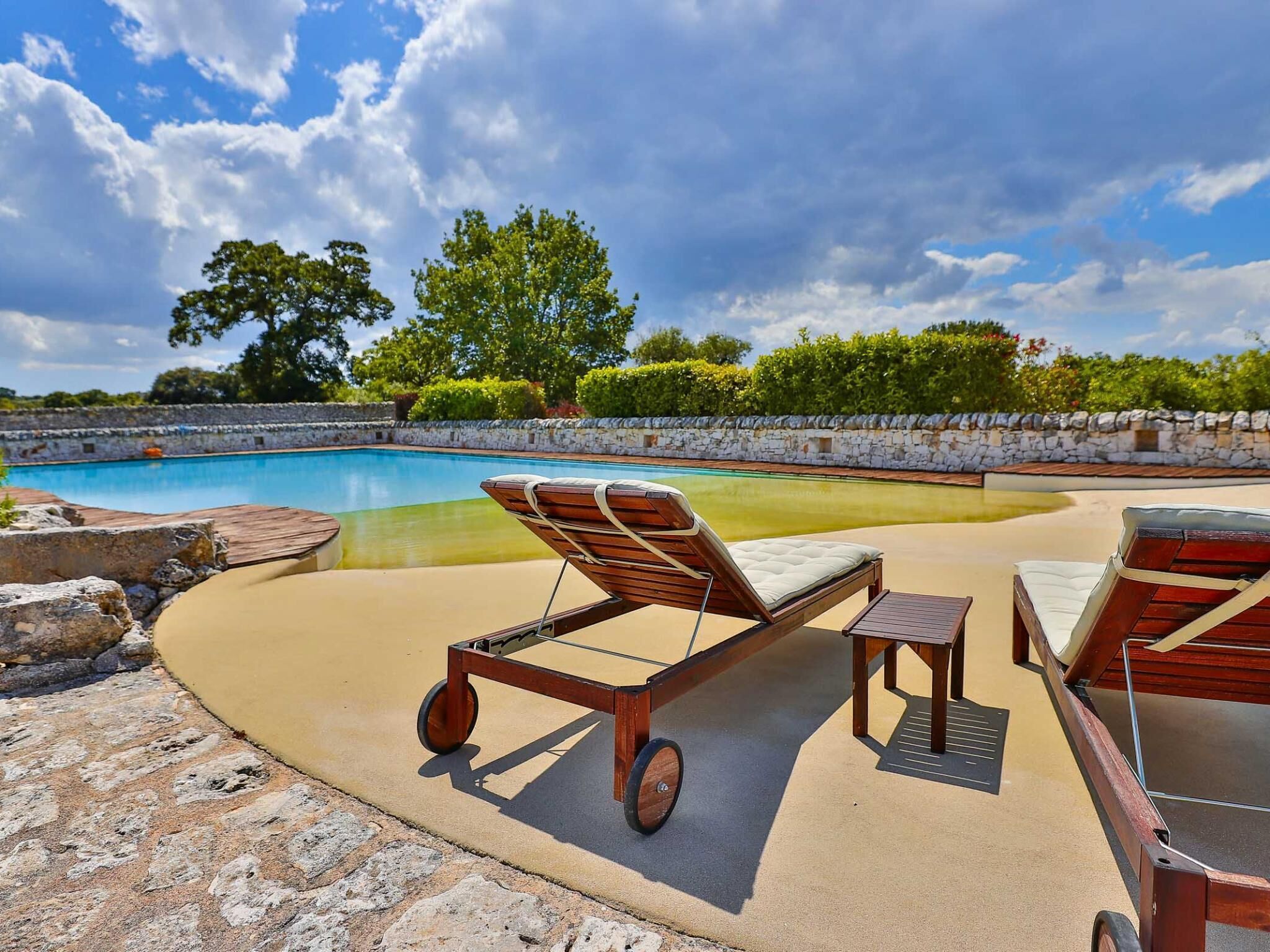 Un espace aménagé au bord de la piscine, avec des chaises longues et un transat, entouré d’un mur de pierre et de verdure.