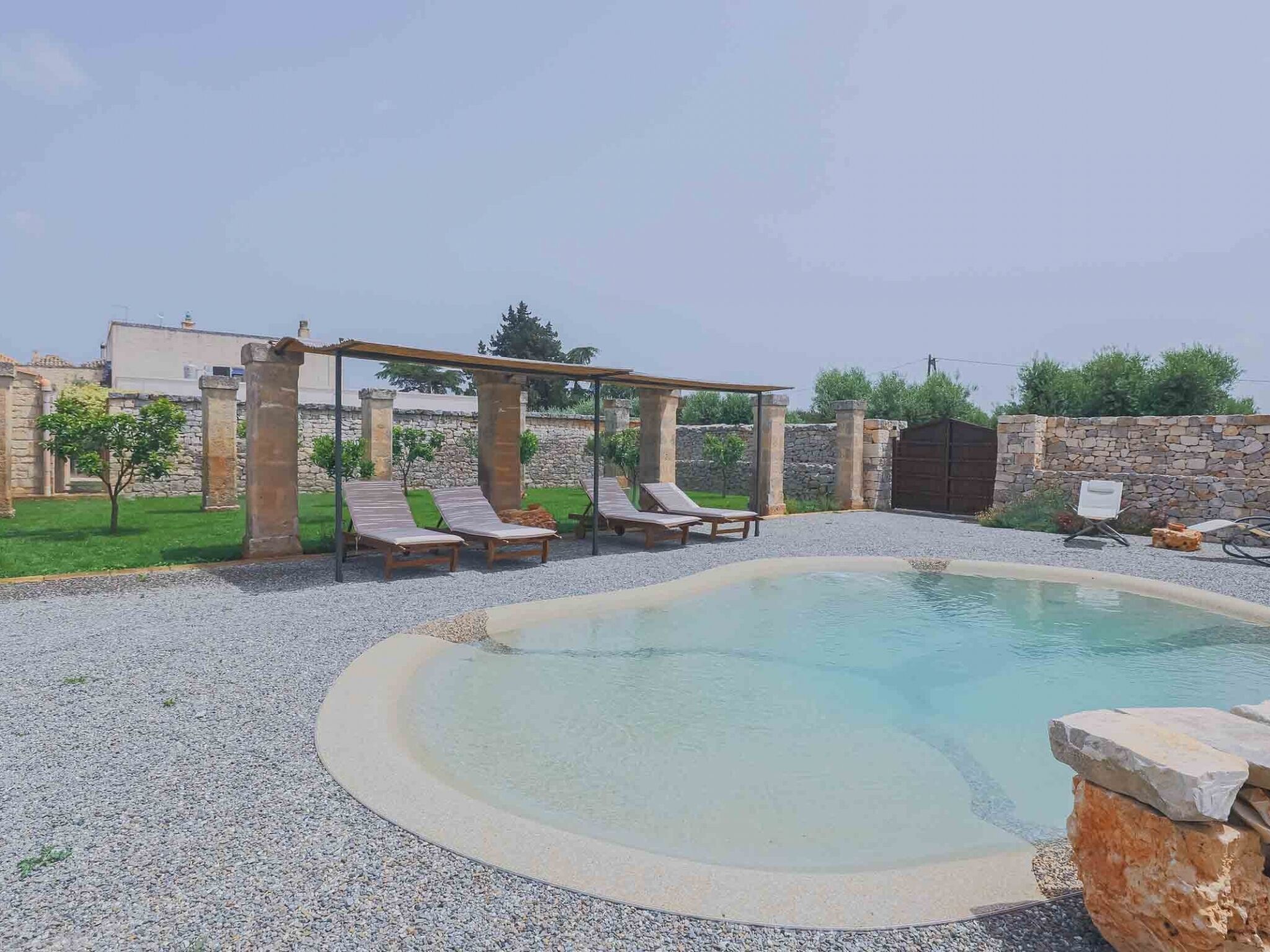 A swimming pool with a pergola and lounge chairs, surrounded by a stone wall and greenery.