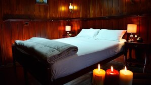 Una cama bien tendida con una colcha blanca y un edredón doblado, dos lámparas de noche y tres velas encendidas sobre el suelo.