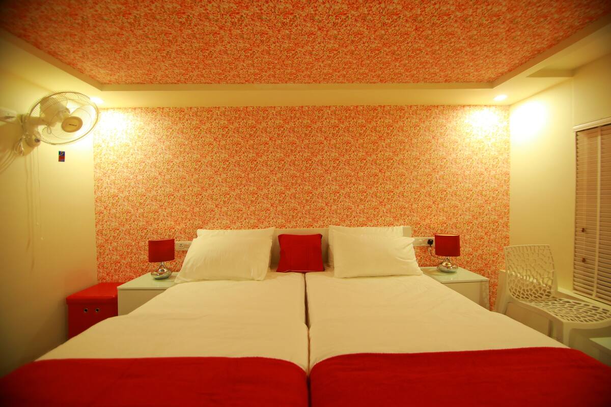 Una cama doble con dos almohadas, una colcha roja, una mesita de noche roja, una lámpara roja, una silla blanca y un ventilador de techo.