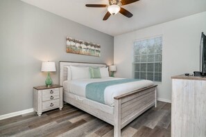 A bedroom with a bed, a nightstand, a ceiling fan, a window with blinds, and a painting on the wall.