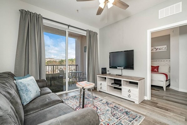 A modern living room with a grey sofa, a flat-screen TV, a wooden entertainment unit, and a balcony with a view.