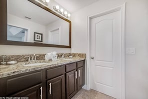 A bathroom with a granite countertop, dark wood cabinets, a white door, and a large mirror with lights.