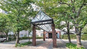 A covered walkway with a glass roof, surrounded by trees and a brick building in the background. - Rupo Kenchoguchi Cozy Hotel (Fukuoka)