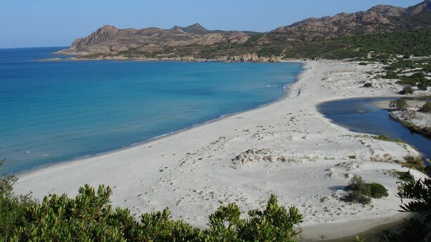 Un paisaje costero con una playa arenosa, aguas azules cristalinas y colinas rocosas al fondo.