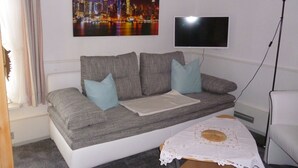 A living room with a grey sofa, two light blue cushions, a white table with a lace tablecloth, and a cityscape painting on the wall.