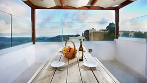 Une table en bois, avec des assiettes, des couverts, une bouteille et un panier de fruits, est placée sur un balcon d’où l’on a une vue dégagée sur le paysage.