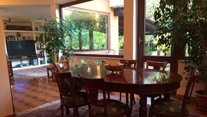 A dining area with a round table and chairs, a television in the background, and large windows with a view of greenery. - Le Querce (Formello)