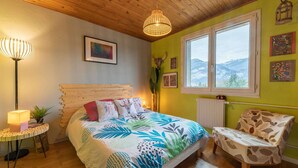A bedroom with a wooden ceiling, a bed with a colorful leaf-patterned bedspread, a side table with a lamp, a chair with a leaf-patterned cushion, and a window with a mountain view. - Back to the 80'S Apartment (Saint-Egrève)