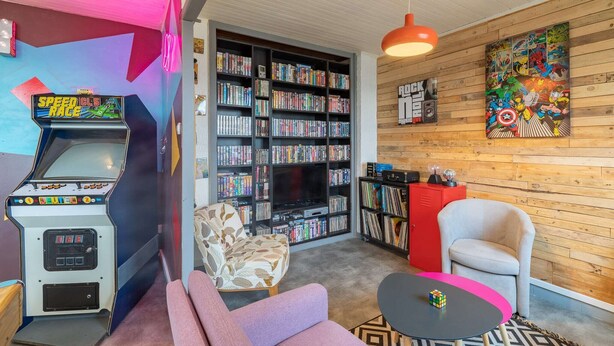 A cozy room with a retro arcade machine, a bookshelf, a comfortable sofa, a small table, and a vibrant wall art piece. - Back to the 80'S Apartment (Saint-Egrève)