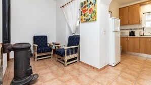 Une pièce chaleureuse avec une cheminée, deux fauteuils, une petite table et un coin cuisine doté de meubles en bois et d’un réfrigérateur.