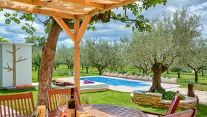 An outdoor patio with a wooden pergola, a round table, and chairs. There is a swimming pool in the background surrounded by trees and a well-maintained lawn. - Casa Oliveto - relax in Nature (Marčana)