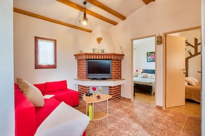 A cozy living room with a red sofa, a small table, a brick fireplace with a TV, and a wooden ceiling beam.