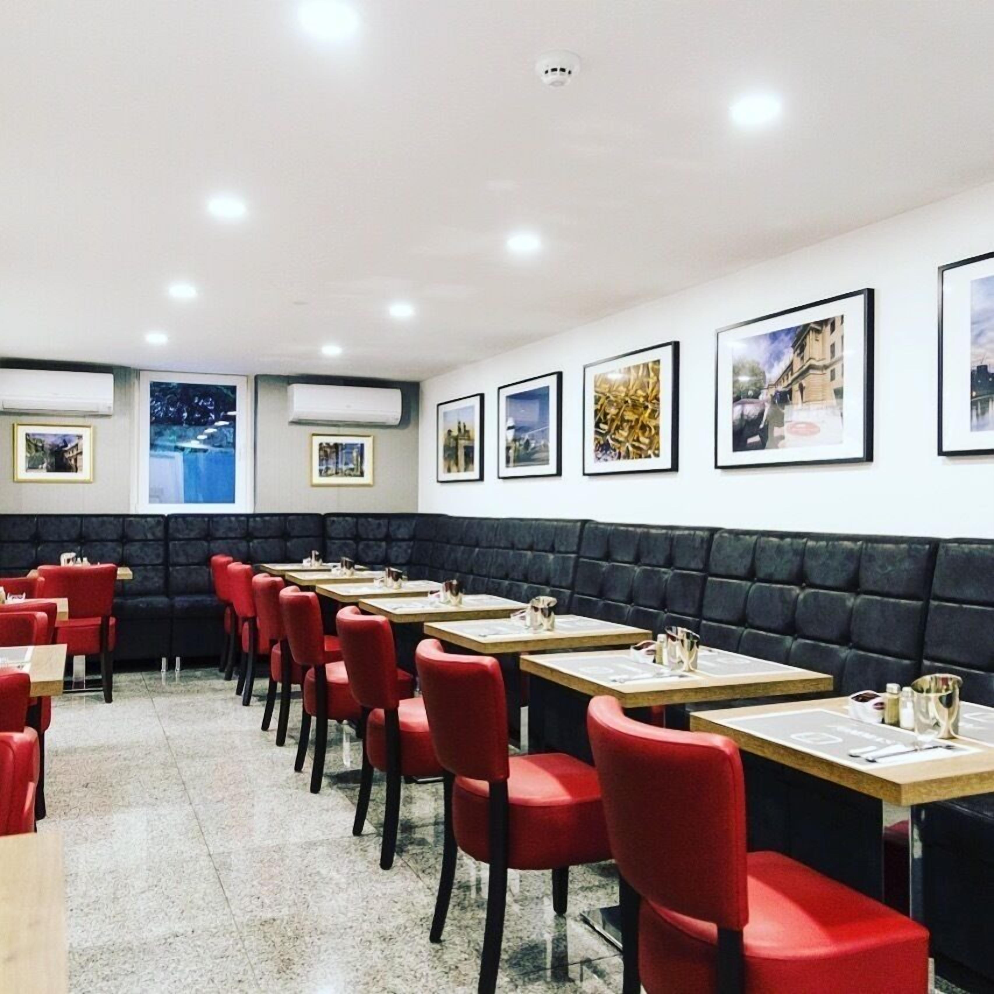 A restaurant with red chairs and tables, a black cushioned bench, and framed pictures on the wall.