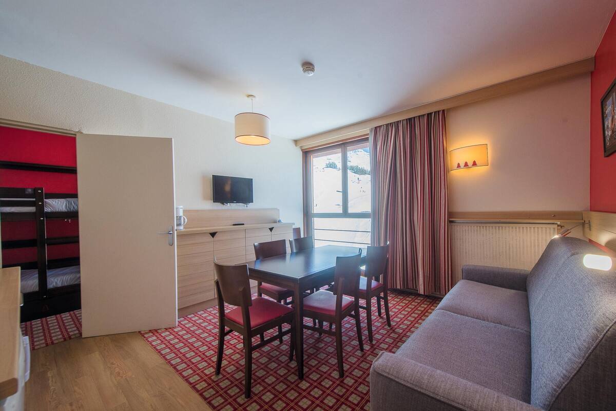 A compact room with a dining table, four chairs, a sofa, a TV, a radiator, and a window with curtains.