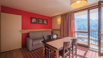 A room with a red wall, a sofa, a table with chairs, a window with curtains, and a view of mountains.