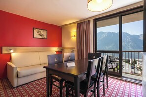 A hotel room with a red wall, a sofa, a dining table with chairs, a large window with a mountain view, and a ceiling light. - Belambra Clubs Orcières 1850 - Le Roc Blanc (Orcieres)