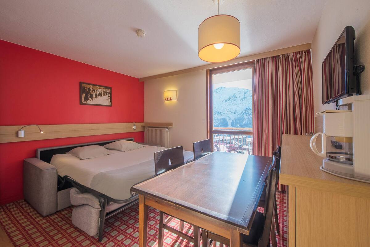 A hotel room with a bed, a dining table, a coffee machine, and a view of mountains.