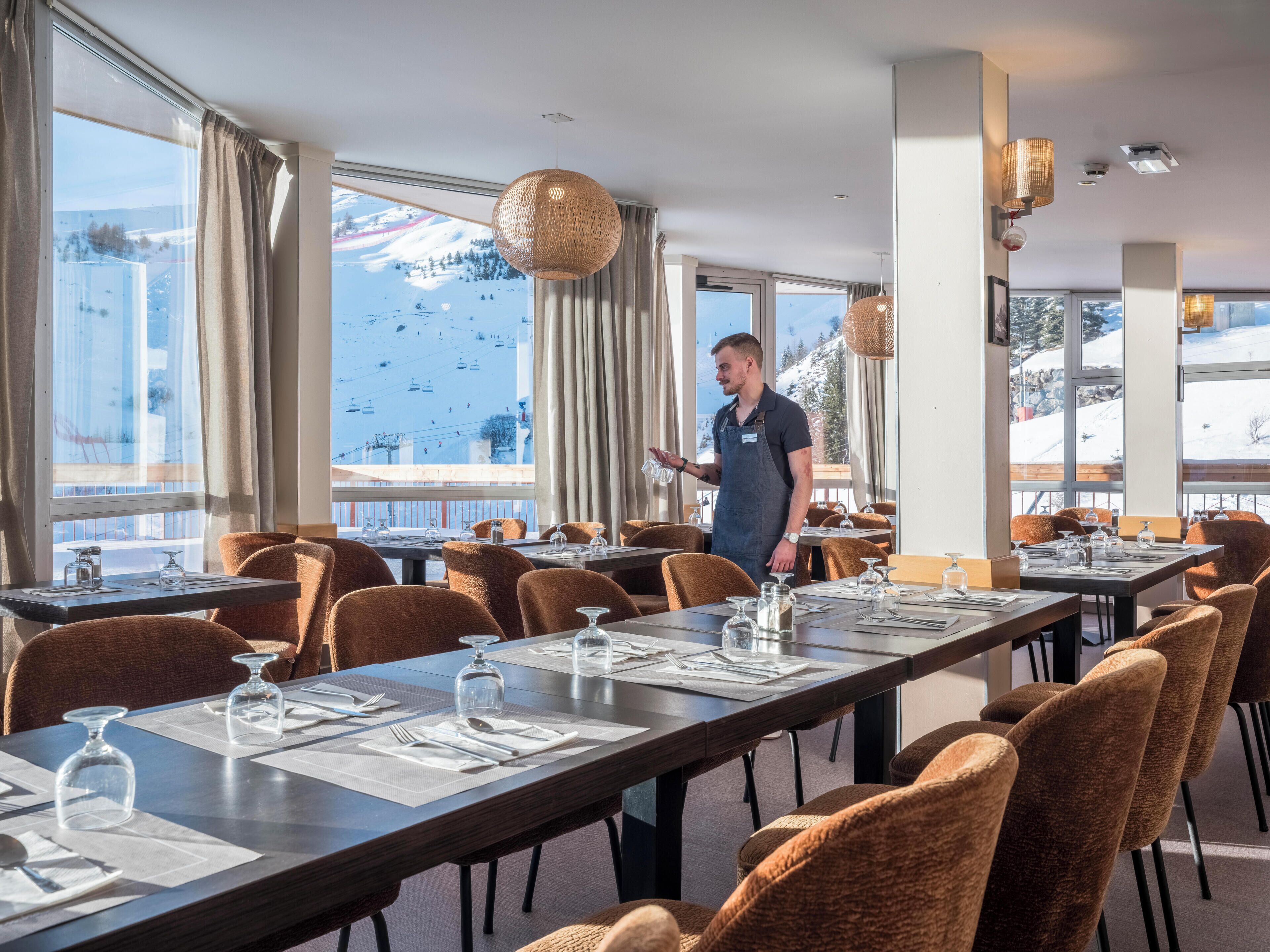 A restaurant with a view of a snowy mountain and ski slope. Tables are set with white tablecloths and glassware.