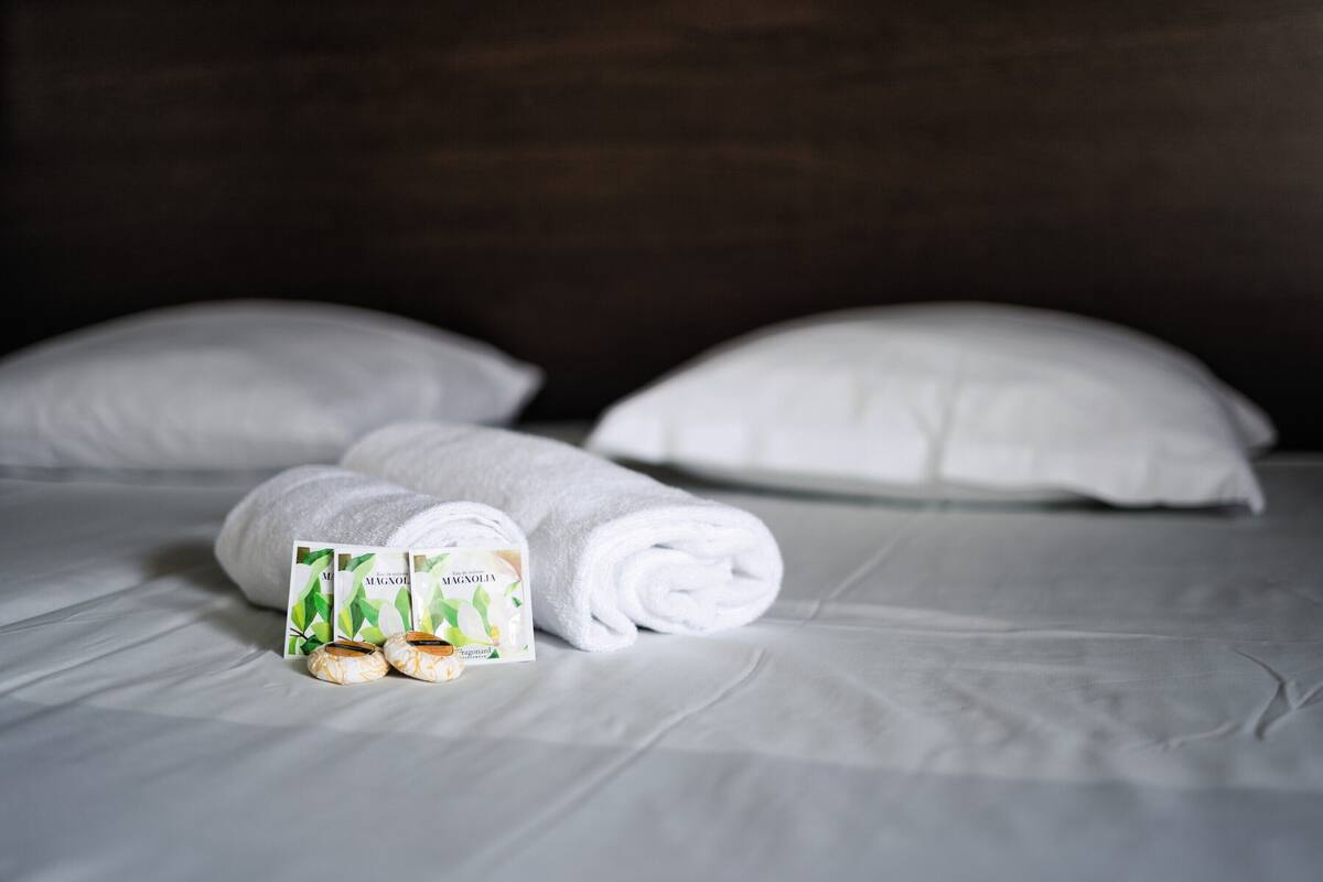 A neatly made bed with white linens, two rolled towels, and a set of toiletries on the bed.