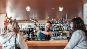 A bar area with a bartender preparing a drink, a customer seated, and various bottles and equipment in the background. - Belambra Clubs Orcières 1850 - Le Roc Blanc (Orcieres)