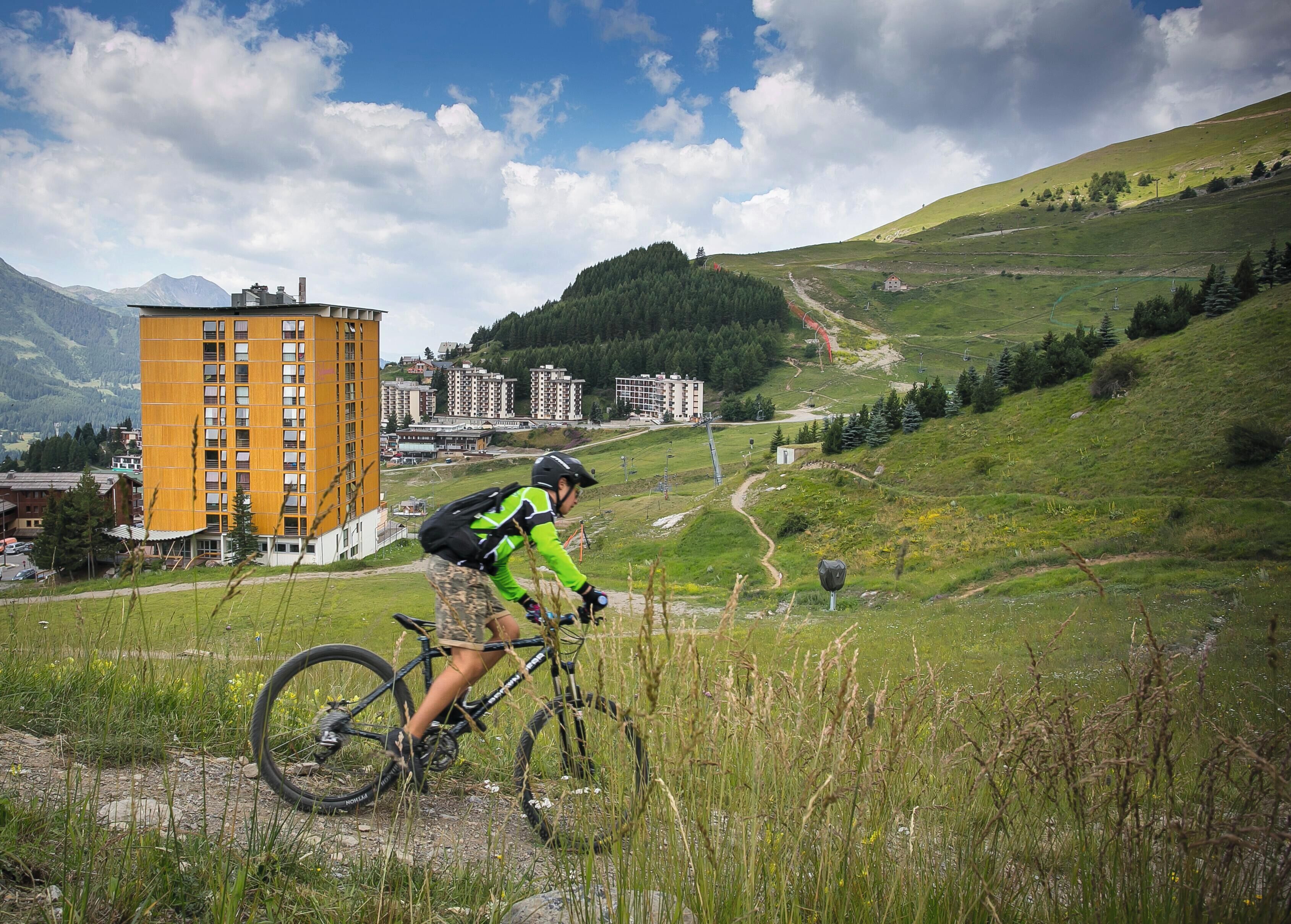 Un ciclista de montaña en un sendero, con edificios al fondo.