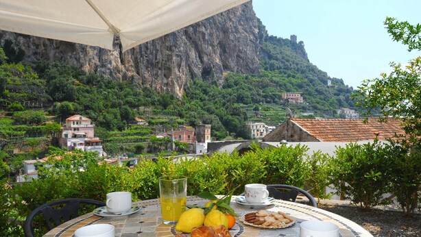 Une table à manger en plein air, dressée pour le petit-déjeuner, entourée d’un paysage pittoresque composé de montagnes et de vignobles en terrasses.