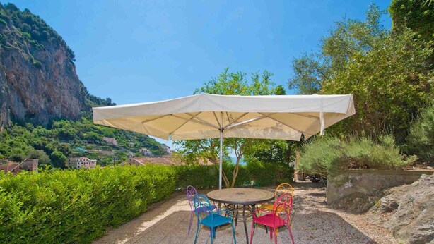 Une terrasse extérieure avec une table ronde et des chaises colorées, protégée par un grand parasol, le tout agrémenté d’un magnifique paysage de colline en arrière-plan.