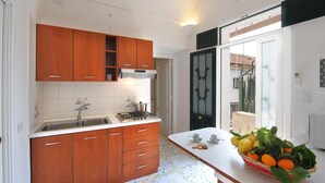 Une cuisine avec des meubles de rangement en bois, une cuisinière à gaz et un évier. Il y a une fenêtre donnant sur un balcon et une porte ornée d’un panneau de verre décoratif.
