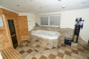 A bathroom with a stone-tiled floor, a wooden door, a large bathtub, a window with blinds, and a water dispenser.