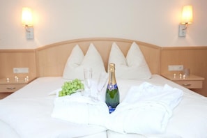 A neatly made bed with white linens, a bottle of champagne, and a bunch of grapes on the bed. There are two bedside tables with lamps and a wooden headboard.